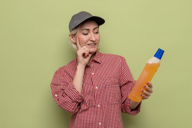 Schoonmaakster in geruit hemd en pet met fles schoonmaakspullen die ernaar kijkt met een glimlach op het gezicht