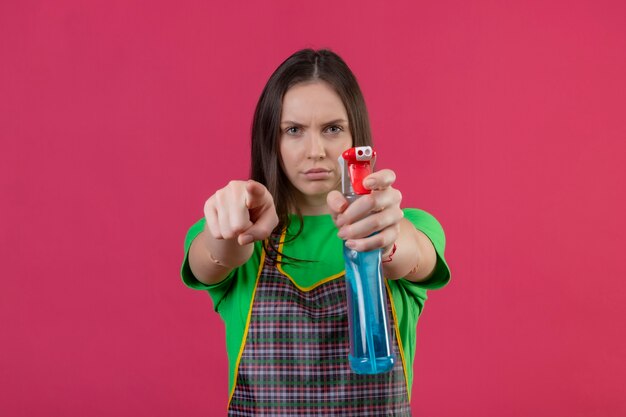 Gratis foto schoonmakende jonge vrouw die eenvormig standhoudend schoonmakende spray draagt die u gebaar op geïsoleerde roze muur toont