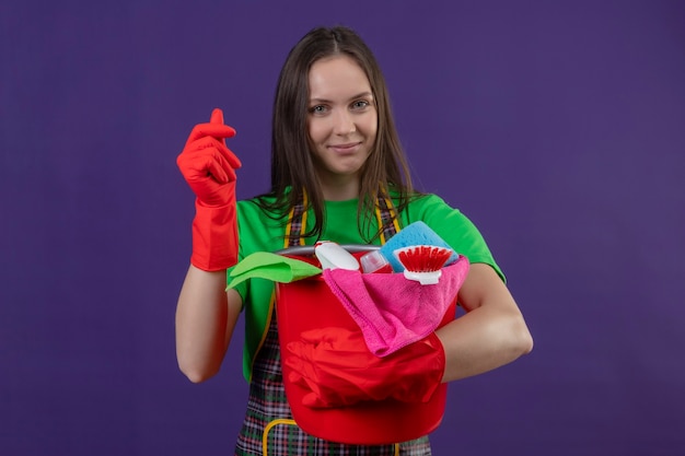 schoonmakende jonge vrouw die uniform in rode handschoenen draagt die schoonmakende hulpmiddelen houdt die contant geldgebaar op geïsoleerde purpere muur tonen