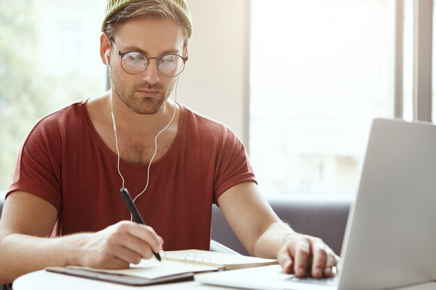 Schot van aantrekkelijke gerichte man in casual kleding, schrijft in notitieblok noodzakelijke informatie