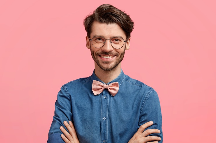 Gratis Foto schot van gelukkige elegante jonge man houdt de handen gekruist, kijkt met vrolijke uitdrukking, heeft een vriendelijke glimlach, draagt een elegant jean-shirt met vlinderdas, geïsoleerd over roze muur. blij mannelijke freelancer