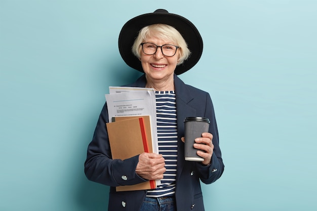 Schot van gelukkige senior ervaren zakenvrouw houdt papieren en portemonnee, drinkt afhaalmaaltijden koffie, blij om succesvol contract te ondertekenen, draagt stijlvolle hoed en jas, vormt binnen. drukke oude werker