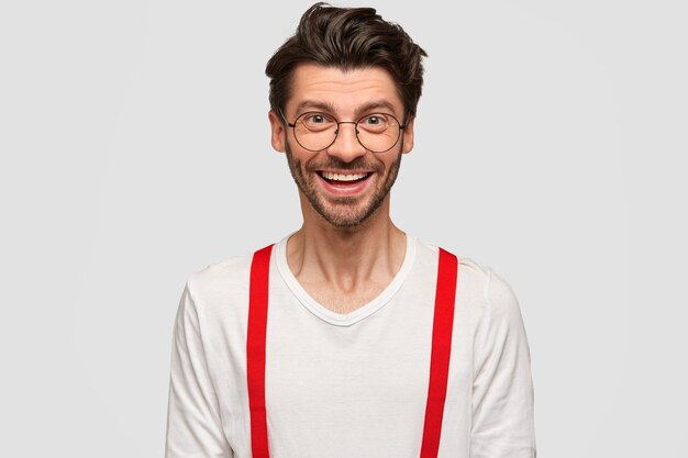Schot van knappe ongeschoren man met een positieve uitdrukking, gekleed in wit overhemd met rode bretels, geïsoleerd op een witte muur. Vrolijke man freelancer in bril verheugt zich op succes in zijn carrière