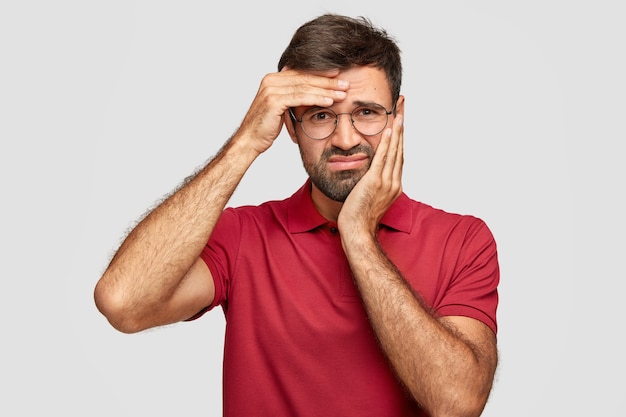 schot van ontevreden bebaarde man heeft kiespijn en hoofdpijn, voelt zich ongelukkig en moe na lang werk, fronst gezicht van ontevredenheid, gekleed in rood t-shirt, geïsoleerd over muur