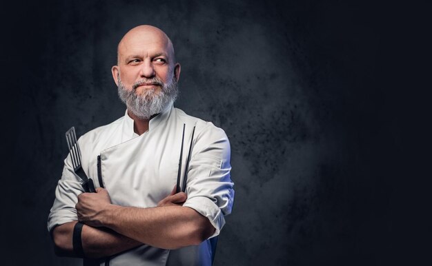 Schot van professionele oude chef-kok gekleed in uniform met keukenspatel en tang.