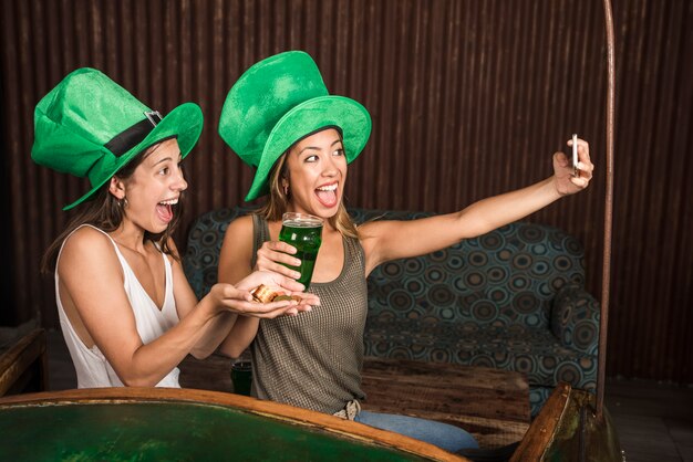 Schreeuwende jonge vrouwen met glas drank en gouden muntstukken die selfie op smartphone nemen