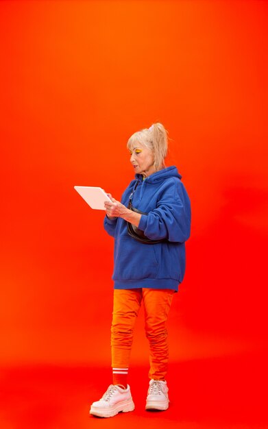 Scrollende tablet. Senior vrouw in ultra trendy kleding geïsoleerd op fel oranje