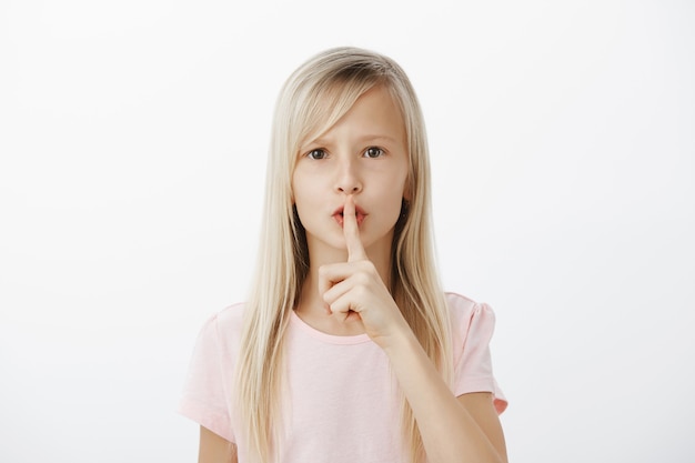 sctrict zelfverzekerd klein blond meisje in casual roze t-shirt, zegt shh terwijl ze een stil gebaar toont met wijsvinger voor de mond, vraagt om stil te zijn terwijl ze voor babybroer zorgt