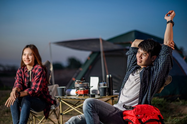 Gratis foto selectieve aandacht handsomman zittend op een stoel en strekkende armen in de buurt van zijn vriendin aan de voorkant van de campingtent, ze glimlachen van geluk en frisheid wanneer ze ontspannen in de natuur.