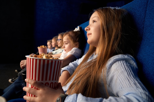 Selectieve aandacht van vrij klein meisje popcorn emmer te houden, zittend met vrienden in comfortabele stoelen in de bioscoop. Kinderen kijken naar tekenfilm of film, plezier maken