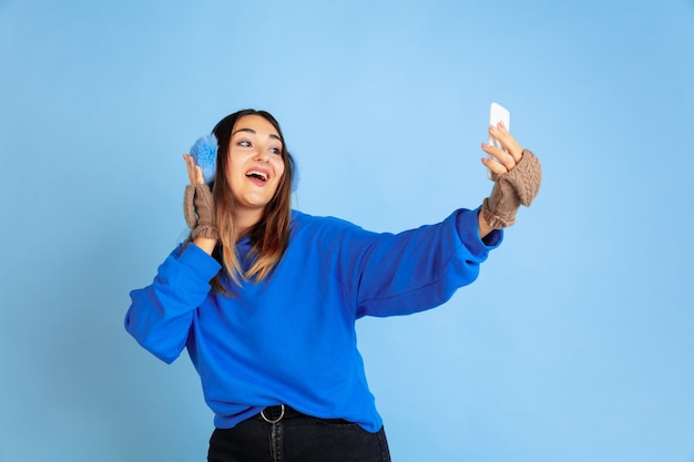 Selfie. Het portret van de Kaukasische vrouw op blauwe studioachtergrond. Mooi vrouwelijk model in warme kleren. Concept van menselijke emoties, gezichtsuitdrukking, verkoop, advertentie. Winterstemming, kersttijd, vakantie.