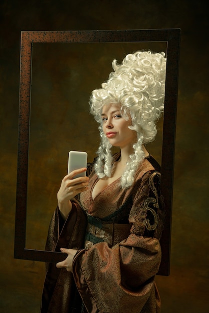 Selfie in spiegel. portret van middeleeuwse vrouw in vintage kleding met houten frame op donkere achtergrond. vrouwelijk model als hertogin, koninklijk persoon. concept vergelijking van tijdperken, modern, mode, schoonheid.