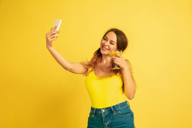 Selfie maken, vlog, glimlachen. Het portret van de Kaukasische vrouw op gele studioachtergrond. Mooi vrouwelijk model. Concept van menselijke emoties, gezichtsuitdrukking, verkoop, advertentie. Zomer, reizen, toevlucht.