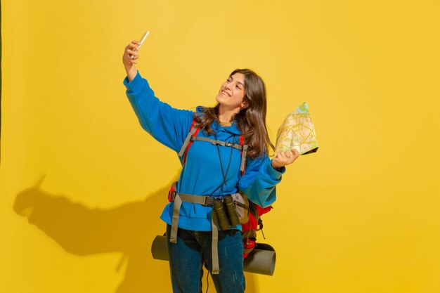 Selfie of vlog maken. Portret van een vrolijk jong Kaukasisch toeristenmeisje met zak en verrekijker dat op gele studioachtergrond wordt geïsoleerd. Voorbereiden op reizen. Resort, menselijke emoties, vakantie.