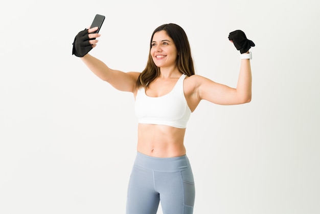 Selfie om te pronken met mijn vooruitgang in de sportschool. Mooie gelukkige vrouw die lacht terwijl ze een foto maakt met haar smartphone en haar sterke biceps laat zien