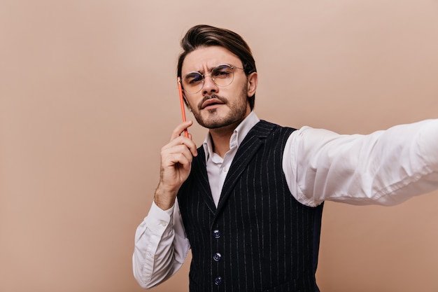 Selfie-portret van denkende man met donkerbruin haar en borstelharen, bril, wit overhemd en klassiek vest, potlood in de buurt van hoofd houden en selfie maken tegen effen beige muur