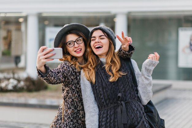 Selfie portret van vrolijke modieuze vrouwen met plezier op zonnige straat in de stad. Stijlvolle look, plezier hebben, reizen met vrienden, glimlachen, echte positieve emoties uitdrukken.
