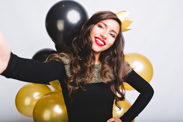 Selfie portret vrolijke vrouw met lang donkerbruin krullend haar, gele kroon, luxe zwarte jurk. Nieuwjaar vieren, verjaardagsfeestje, plezier maken met gouden en zwarte ballonnen.