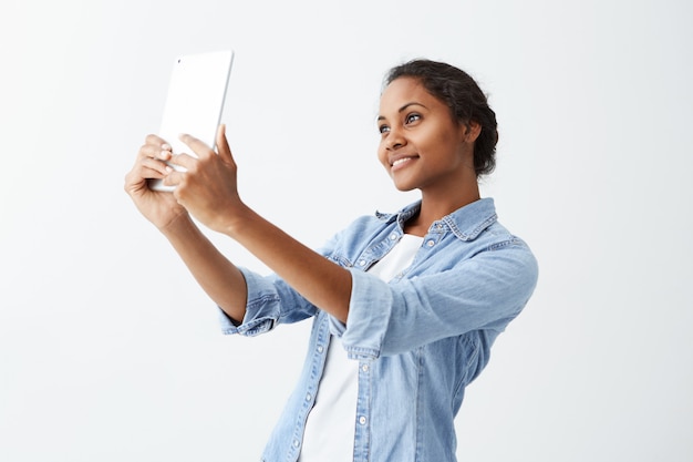 Selfie tijd. Vrolijke jonge aantrekkelijke Afro-Amerikaanse vrouw met donker haar in blauw shirt maken selfie, tablet in haar handen houden. Het donkere beautidul meisje van de donkere huid het stellen voor selfie op witte muur
