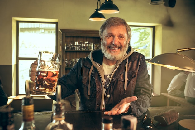 Senior bebaarde man alcohol drinken in pub en kijken naar een sportprogramma op Tv. Genieten van mijn favoriete krieltjes en bier. Man met mok bier aan tafel zitten. Voetbal- of sportfan. Menselijke emoties concept