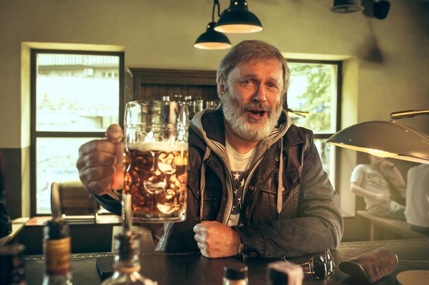 Senior bebaarde man alcohol drinken in pub en kijken naar een sportprogramma op Tv. Genieten van mijn favoriete krieltjes en bier. Man met mok bier aan tafel zitten.