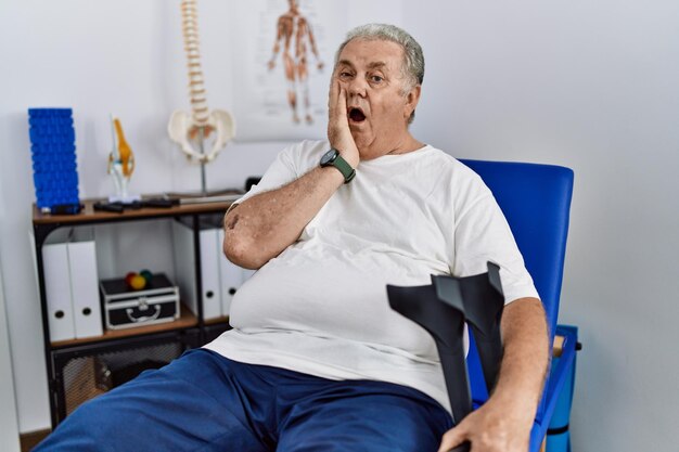 Senior blanke man in de fysiotherapiekliniek met krukken bang en geschokt, verrassing en verbaasde uitdrukking met de handen op het gezicht