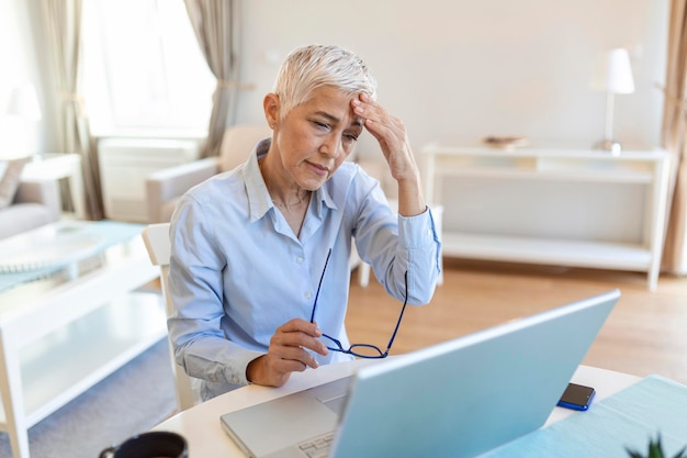 Senior gefrustreerde vrouw die vanuit een thuiskantoor voor een laptop werkt met hoofdpijn rijpe vrouw met een pijnlijke gezichtsuitdrukking