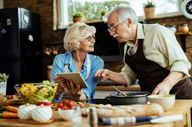 Senior koppel bereidt lunch voor terwijl ze het recept volgen op touchpad in de keuken