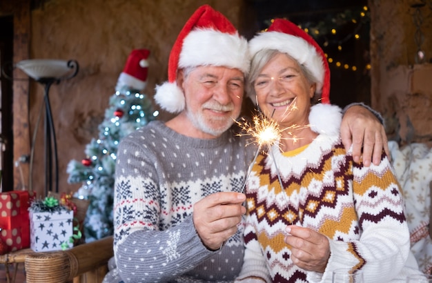 Senior lachende paar in kerstmutsen knuffelen thuis met kerstmis met wonderkaarsen. kerstboom, cadeautjes en decoraties op de achtergrond