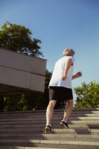 Senior man als hardloper met armband of fitnesstracker op straat in de stad. Kaukasisch mannelijk model dat joggen en cardiotrainingen beoefent in de zomerochtend. Gezonde levensstijl, sport, activiteitenconcept.