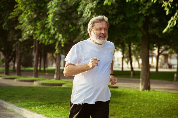 Senior man als hardloper met armband of fitnesstracker op straat in de stad. Kaukasisch mannelijk model joggen en cardio-trainingen in de zomerochtend. Gezonde levensstijl, sport, activiteitenconcept.
