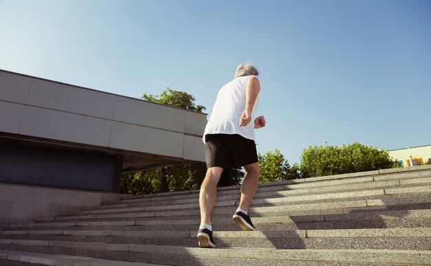 Senior man als hardloper met armband of fitnesstracker op straat in de stad. Kaukasisch mannelijk model joggen en cardio-trainingen in de zomerochtend. Gezonde levensstijl, sport, activiteitenconcept.