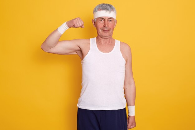 Senior man binnen dragen van sportkleding en hoofdband, staan met een hand omhoog en tonen zijn biceps, gefotografeerd na het doen van fysieke oefeningen. Gezonde levensstijl concept.