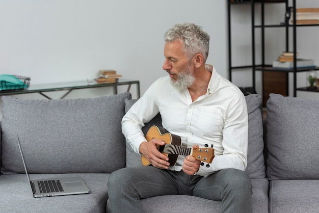 Gratis foto senior man die thuis ukelele-lessen studeert op laptop