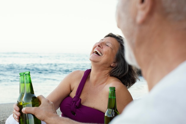 Senior man en vrouw lachen op het strand terwijl ze bier drinken