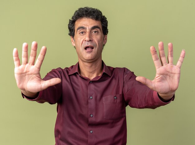 Senior man in paars shirt kijkend naar camera bang om stopgebaar te maken met handen die handen uitstrekken boven groen