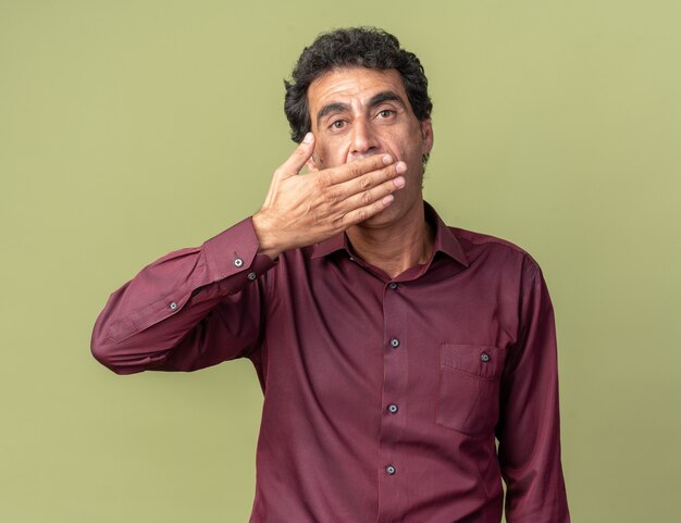 Senior man in paars shirt kijkend naar camera die geschokt is en mond bedekt met hand die over groene achtergrond staat