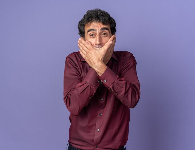 Senior man in paars shirt kijkend naar camera die geschokt is en mond bedekt met handen die over blauwe achtergrond staan