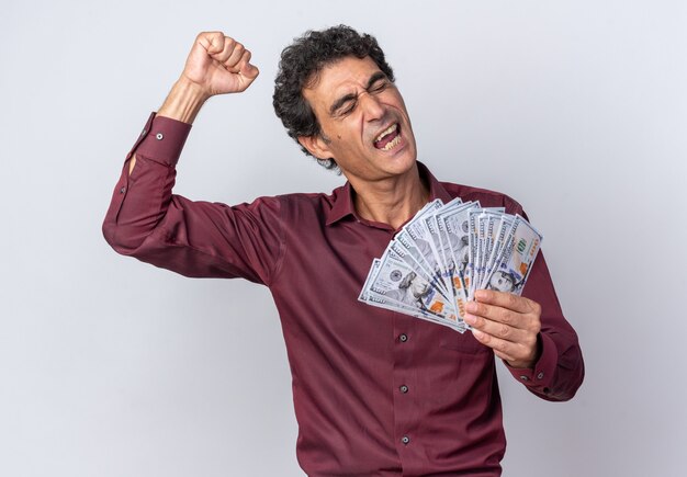 Senior man in paars shirt met contant geld blij en opgewonden, gebalde vuist op een witte achtergrond