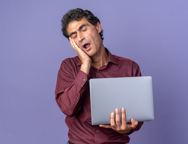 Senior man in paars shirt met laptop die er moe en verveeld uitziet geeuwen