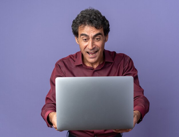 Senior man in paars shirt met laptop die er verbaasd en verrast naar kijkt