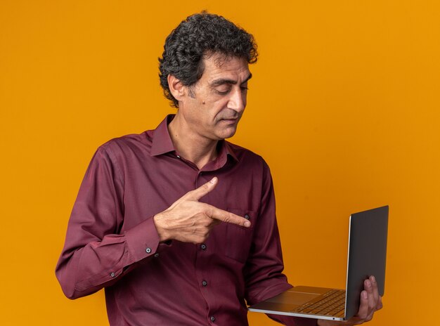 Senior man in paars shirt met laptop die met wijsvinger erop wijst en er zelfverzekerd uitziet als hij over oranje staat