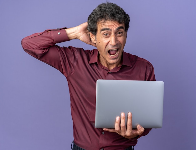 Senior man in paars shirt met laptop die schreeuwt en verbaasd en verrast over een blauwe achtergrond staat