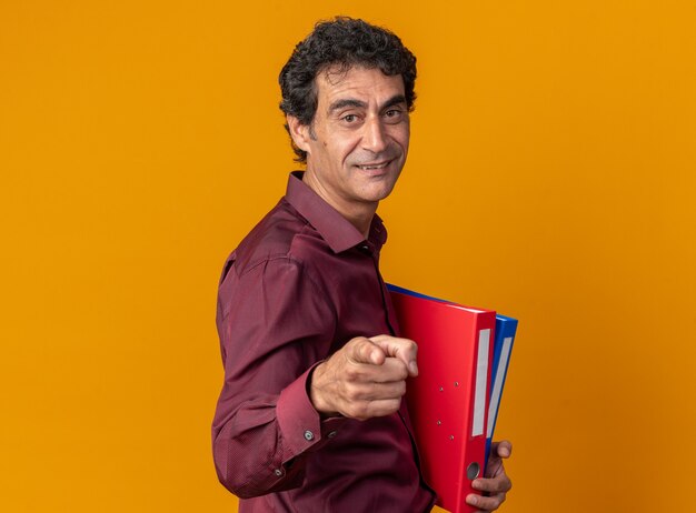 Senior man in paars shirt met mappen kijkend naar camera wijzend met wijsvinger naar camera glimlachend met blij gezicht over oranje achtergrond