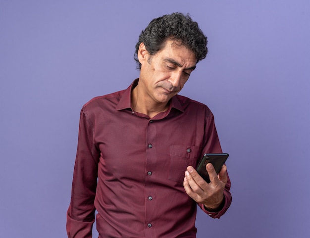 Senior man in paars shirt met smartphone die ernaar kijkt met een serieus gezicht over blauw