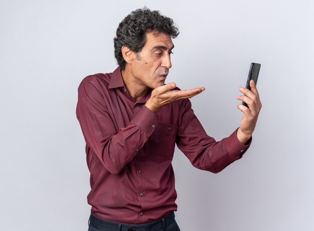 Senior man in paars shirt met smartphone die naar het scherm kijkt en een kus blaast terwijl hij over wit staat