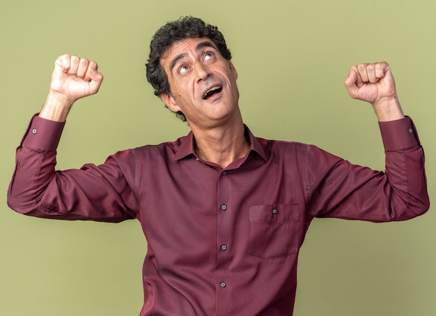 Senior man in paars shirt met vuisten blij en opgewonden staande over groene achtergrond