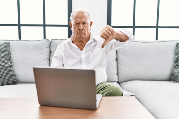 Senior man met behulp van laptop thuis zittend op de bank kijken ongelukkig en boos tonen van afwijzing en negatief met duim omlaag gebaar slechte uitdrukking