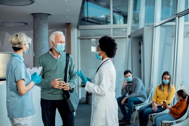 Senior man met beschermend gezichtsmasker in gesprek met artsen in de gang van het ziekenhuis