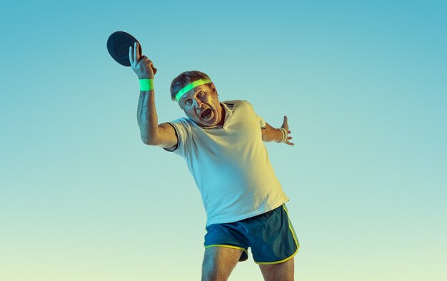 Senior man tafeltennis spelen op verloopmuur in neonlicht. Kaukasisch mannelijk model in uitstekende vorm blijft actief, sportief. Concept van sport, activiteit, beweging, welzijn, gezonde levensstijl.